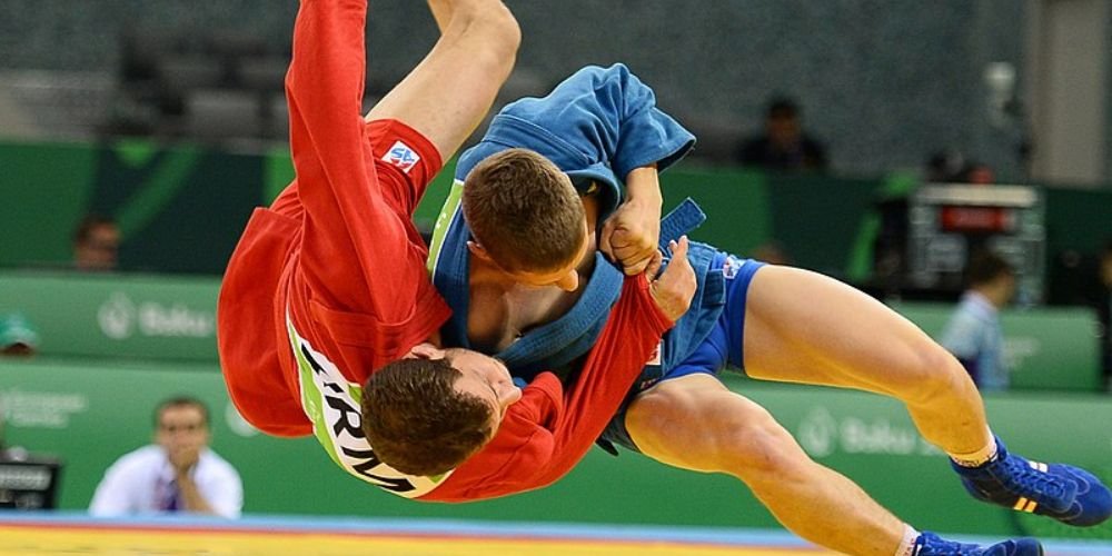 Dos luchadores de Sambo masculino compiten intensamente sobre el tatami. Uno de ellos, vestido con chaqueta roja, intenta proyectar a su oponente, que lleva uniforme azul, en una técnica típica del Sambo deportivo. Ambos muestran concentración y técnica en un combate oficial